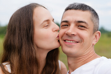 A woman with long brown hair kisses a smiling man on the cheek, with a blurred natural background suggesting an outdoor setting.