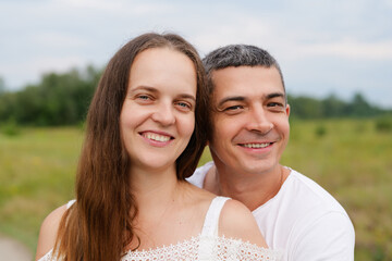A happy couple smiles directly at the camera, with the woman in the foreground and the man behind her, against a soft, blurred green outdoor background.