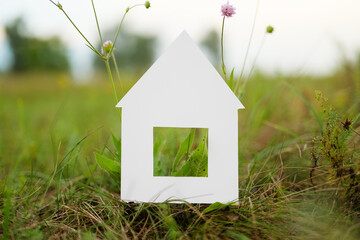 A white paper cutout of a house stands in green grass and wildflowers, symbolizing dreams, nature, and the hope of building a home in harmony with the land.
