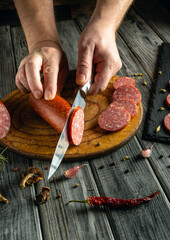 Hands are skillfully cutting through a rolled sausage on a wooden board. Surrounding ingredients...