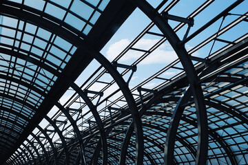 Fototapeta premium Modern industrial steel and glass structure. Geometric roof pattern with curved metal beams and transparent panels. Urban architecture design. Blue sky visible through construction.