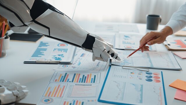 This image captures a robotic arm working alongside a human hand over various business charts and graphs in a bright office environment, showcasing modern automation.Noogenesis