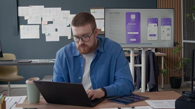 Portrait of young male UX designer typing on laptop creating UI prototype for mobile app then turning to camera with smile while working at office, slow motion