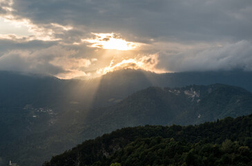 Mystical light breaking through the clouds over the mountains near Illegio, casting golden rays on the forested hills below.