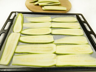 Slices of fresh zucchini are  on a baking parchment on a baking sheet before a roasting of the vegetables in the oven.
