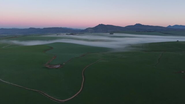 Aerial view of expansive green fields meet a blanket of ethereal fog, with distant mountains painted under a soft pink and blue sky, Caledon, Western Cape, South Africa.