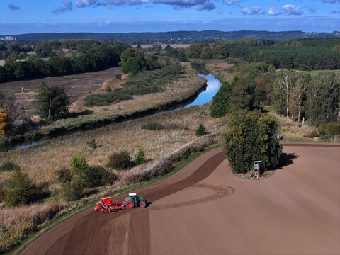 Aerial view of a tractor tilling the soil near a winding waterway, contrasting with the lush greenery of the surrounding landscape, Bralitz, Bad Freienwalde, Brandenburg, Germany.