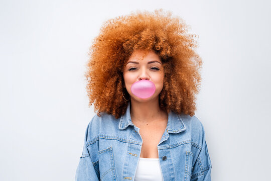 Playful latin young woman feeling positive emotion showing satisfied expression, having fun with bubblegum at white wall. Isolated portrait of smiling ginger teenager girl blowing pink chewing gum. 
