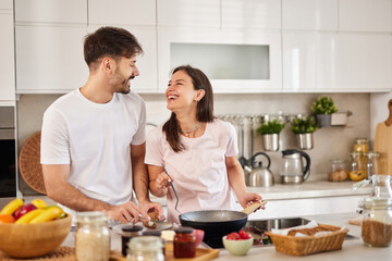 Couple stands close in a well-lit kitchen, laughing joyfully as they cook together. Ingredients and...