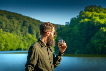 Bearded man holding a cup, enjoying morning coffee outdoors by a lake