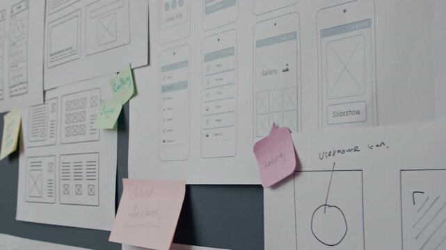 Close up of printed wireframes of mobile app hanging on wall covered with colorful stickers with handwritten notes at UX designers office