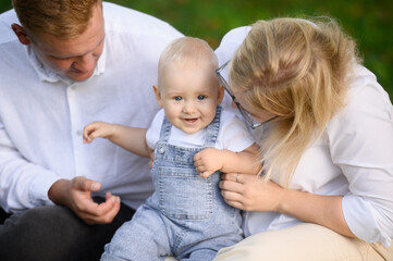 Happy baby sitting on hands of young parents in park on summer weekend, parents lovingly looking at baby, tickling him, close-up. Family values, outdoor recreation.