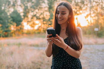 Smiling young woman with long hair using a smartphone outdoors at sunset, wearing a black polka dot...