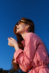 Joyful young woman in pink outfit smiling against a blue sky background, embodying happiness and...