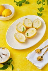 Dessert, lemon posset or citrus mousse in lemon peel halves decorated with lemon zest and mint on a white oval dish on a yellow background. Citrus desserts.