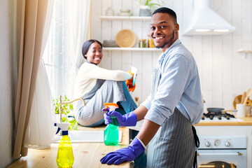 Positive black guy using detergent for house cleanup, his girlfriend sitting on kitchen table, copy...