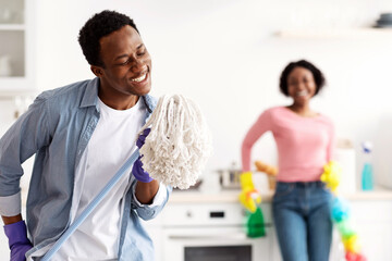Joyful black guy using mop as microphone, singing while cleaning kitchen with his beautiful...