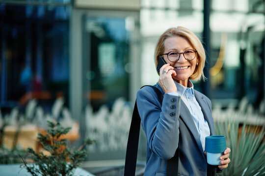 Portrait of a middle aged mature businesswoman woman using a smartphone mobile phone and holding a cup of coffee walking down the street, surrounded by moder corporate office buildings