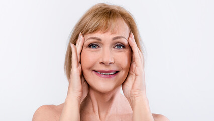 Senior woman touching her face and smiling at camera, enjoying results of her anti-aging skin care...