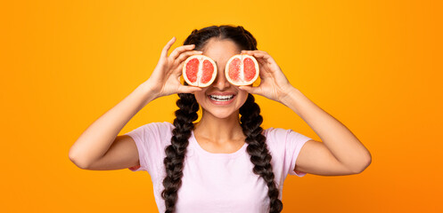 Having Fun, Summer Mood. Funny playful young lady holding halves of citrus juicy grapefruit against her eyes, covering them isolated over bright yellow studio background. Vitamins And Healthy Diet