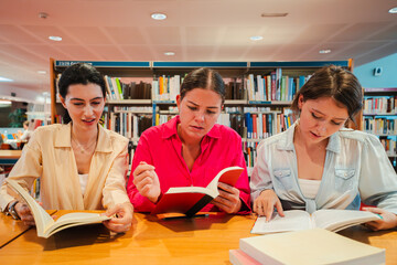 Three dedicated young women are deeply immersed in their reading materials, demonstrating focus and collaboration while studying together with books in a library environment for academic purposes.