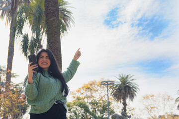 Young Venezuelan woman in the square with her phone in her hand and pointing her finger at the sky