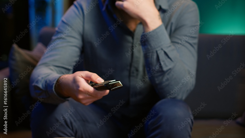 Poster close up of man in dimly lit apartment using remote control to browse though movies on streaming ser