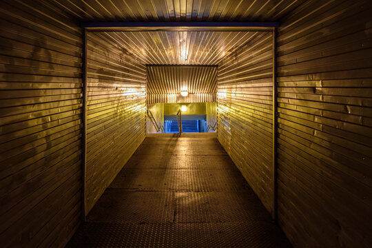 Illuminated metal pedestrian underpass tunnel at night with yellow and blue artificial lighting. Urban subway corridor with metal walls, stairs, and perspective lines. Empty, industrial, moody scene.