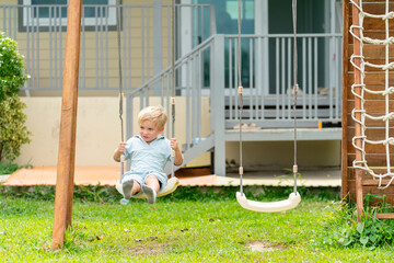 Happy little child boy swinging on playground. Children enjoy and fun outdoor active lifestyle leisure activity playing on swing at backyard. Kindergarten preschool kid playing outside classroom.