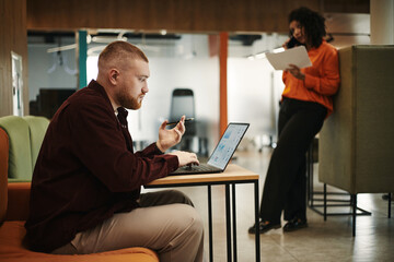 Caucasian young man sitting at desk using laptop and holding smartphone, Black woman standing in...