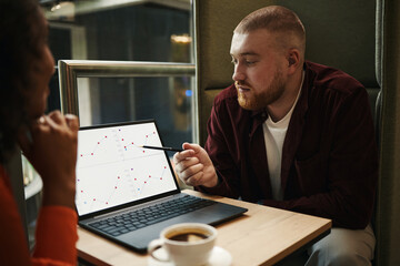 Caucasian young man explaining financial charts on laptop screen to Black woman in business...