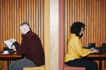 Caucasian young man reading notebook while sitting at table next to Black woman working on laptop,...