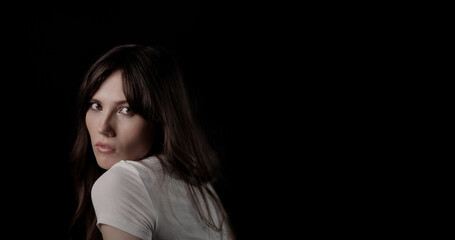 Obraz premium Beautiful young woman with long brown hair wearing white t shirt looking over her shoulder on black background