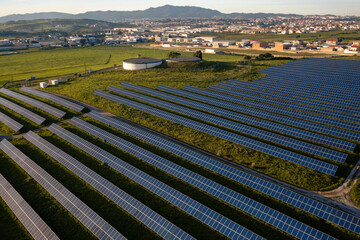 Rows of solar panels stretch across a vibrant green field, harnessing sunlight as the sun sets. Nearby towns contribute to the serene landscape. Scene showcases sustainable energy solutions.