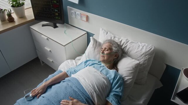 High angle shot of senior female patient with pulsometer and oxygen tubes sleeping in hospital bed. Huge monitor showing her heart rate and pulse