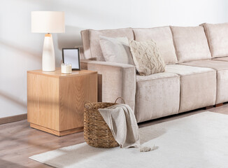 Living Space with Beige Upholstered Sofa with Textured Pillows, Wooden Side Table Hosting Ceramic Lamp, Picture Frame, Woven Basket with Throw Blanket, Set on Light Wooden Flooring and White Rug.