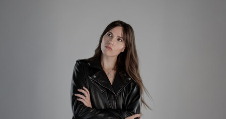 Young woman with long brown hair wearing black leather jacket is posing with arms crossed on gray...