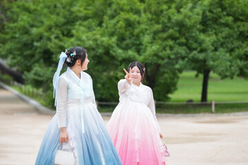 Two Korean women in their twenties walk together through a lush green park wearing cute pink and...