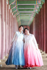 Fototapeta premium Two Korean women in their twenties take a commemorative photo among the columned corridors of Gyeongbokgung Palace, wearing cute pink and sky-blue hanbok beneath a bright summer sky. Seoul, June.