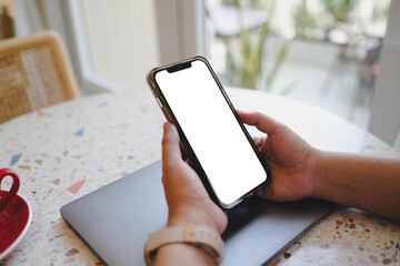 A person holding a smartphone with a blank white screen in a cafe, with a laptop and a red coffee cup on the table. A versatile mobile app mockup.