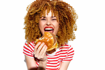 Smiling young woman with curly hair enjoying a hamburger, wearing a striped shirt, capturing joy and indulgence in a bright, cheerful setting