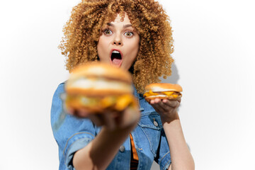 Surprised woman with curly hair holding burgers, displaying excitement for fast food, wearing denim jacket, white background emphasizing colors and seasonal appeal