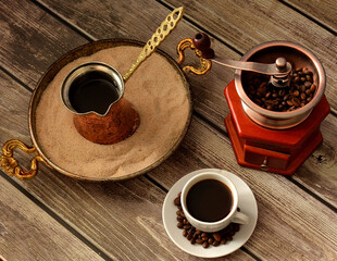 Cup of black coffee on a saucer with roasted beans, tray with sand and cezve and manual coffee grinder on a wooden table.