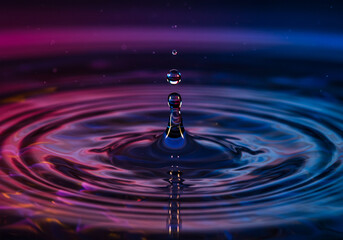 A water drop creates perfect concentric ripples on a surface, illuminated by a vibrant, multi-colored light