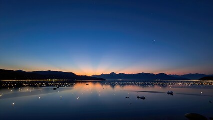Serene twilight scene showcases tranquil lake reflecting vibrant hues of sunset with distant mountains and shimmering lights. Glaciers