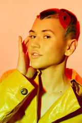 Woman with colorful hair and yellow coat posing confidently against warm peach background,...