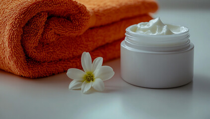White round plastic jars filled with white cream, placed on a table next to two bright orange towels and a small delicate flower, set against a clean white background,