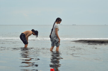 Two teen girls walk carefully across a shallow estuary with pants lifted, surrounded by waves, wet...