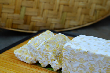A block of tempeh, partially sliced, sits on a wooden cutting board with a woven bamboo mat in the background.