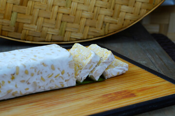 A block of tempeh, partially sliced, sits on a wooden cutting board with a woven bamboo mat in the background.